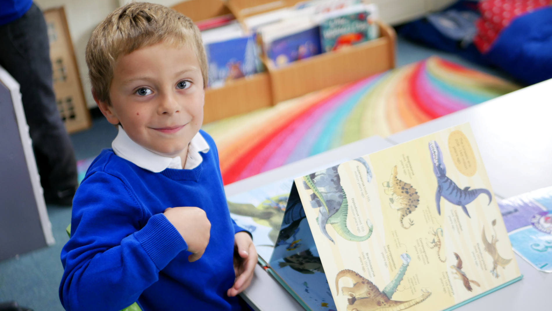 pupil smiling at the camera as we take a picture of him reading a dinosaur book in english at horton kirby primary school