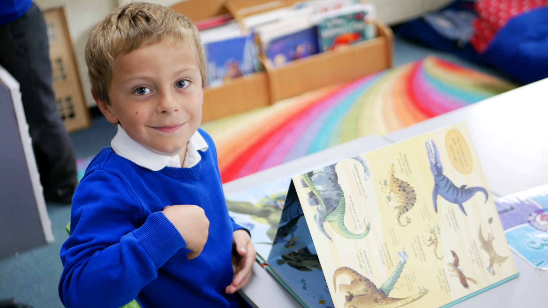 dinosaur book pupil smiling at the camera as we take a picture of him reading a dinosaur book in english at horton kirby primary school